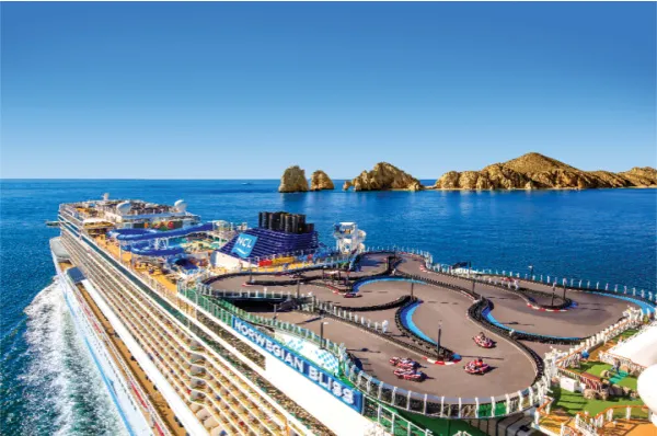 Luftaufnahme des Kreuzfahrtschiffes Norwegian Bliss auf dem blauen Ozean vor der Felsenküste von Cabo San Lucas, mit gut sichtbarer Go-Kart-Rennstrecke auf dem obersten Deck.