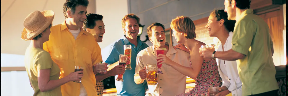 Fröhliche Gruppe von Kreuzfahrtgästen, die mit verschiedenen Drinks wie Bier und Cocktails an einer Bar an Deck stehen, gemeinsam lachen und die entspannte Atmosphäre genießen.
