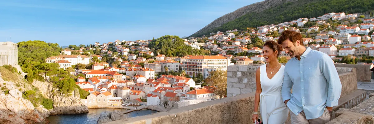 Ein Paar spaziert entspannt über eine alte Festungsmauer mit Blick auf die sonnigen, roten Ziegeldächer der Altstadt von Dubrovnik und grüne Berghänge im Hintergrund.