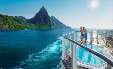 Rückansicht eines Paares in Badekleidung, das an der Reling des Sonnendecks eines Kreuzfahrtschiffes steht und auf das tiefblaue Meer sowie die grünen Berge einer tropischen Insel blickt, direkt neben einem eingelassenen Whirlpool.