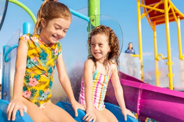 Poolbereich auf der Legend of the Seas Zwei Mädchen in Badeanzügen sitzen auf einer Wasserrutsche und lachen miteinander.