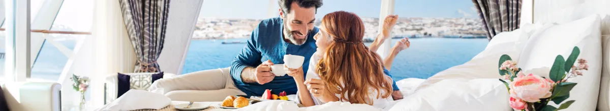 Ein Paar genießt Ihre NCL-Kreuzfahrt in einer The Haven Suite auf der Norwegian Star. Sie liegen mit Kaffee auf dem Bett.