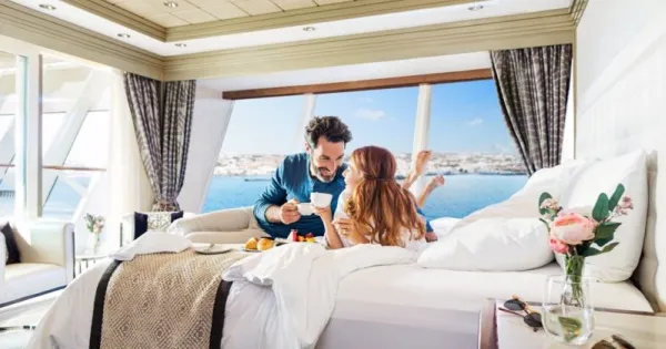 Ein Paar liegt auf dem Beet in einer Balkonkabine und genießt eine Tasse Kaffee, im Hintergrund Meer und blauer Himmel. Die Kabine ist in hellen Farben gestaltet und mit frischen Blumen in einer Vase dekoriert.