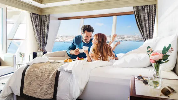 Kabinen Norwegian Cruise Line Ein Paar liegt auf dem Beet in einer Balkonkabine und genießt eine Tasse Kaffee, im Hintergrund Meer und blauer Himmel. Die Kabine ist in hellen Farben gestaltet und mit frischen Blumen in einer Vase dekoriert.