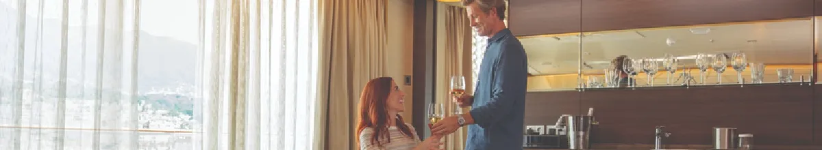 Ein glückliches Pärchen in einer The Haven Suite mit Panoramafenstern mit Gardinen und einer Bar im Hintergrund.