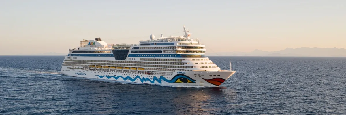 Genießen Sie die Weite des Ozeans bei zauberhaften Transreisen mit AIDAblu & Co. Seitenansicht von AIDAblu an einem sonnigen Tag auf dem Meer. Im Hintergrund sind schemenhaft die Umrisse einer Bergkette zu erkennen.