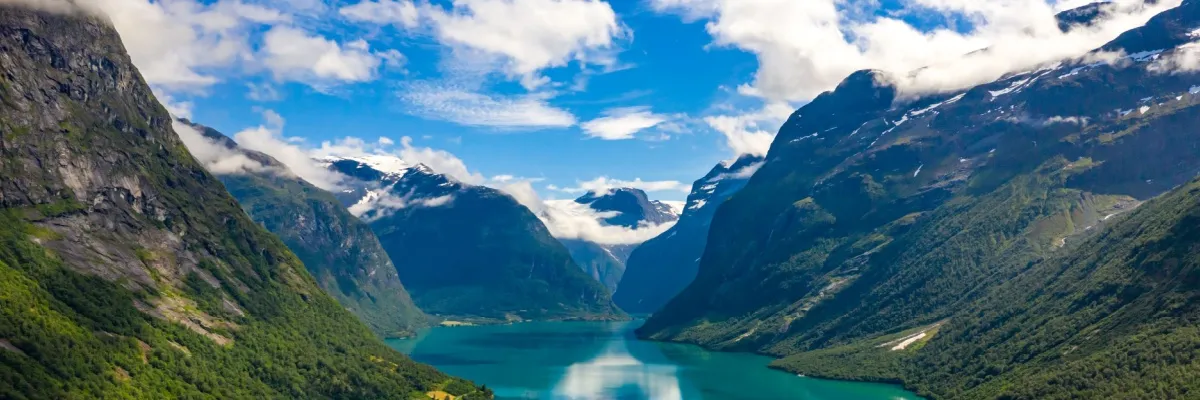 AIDA Travel Tuesday: Im Frühling nach Nordeuropa Geirangerfjord in Norwegen: Türkisblaues Wasser zwischen steilen, grün bewachsenen Hängen. Darüber ein leicht bewölkter blauer Himmel.