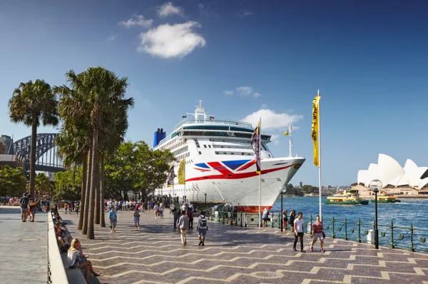 Frontalansicht der Aurora von P&O Cruises an einem Kai in Sydney, im Hintergrund ist die Oper und im Vordergrund eine Promenade mit Menschen.