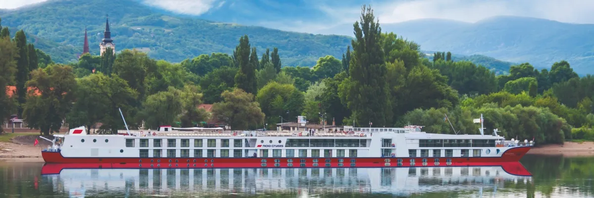 Seitenansicht der MS BOLERO auf der Donau. Im Hintergrund sind Bäume, bewaldete Hügel sowie die Spitzen zweier Kirchtürme zu sehen.
