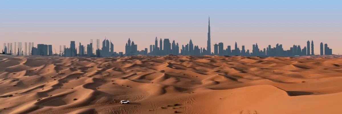Eine weite Wüste erstreckt sich vor der Skyline Dubais. Ein Geländewagen fährt durch die Dünen.