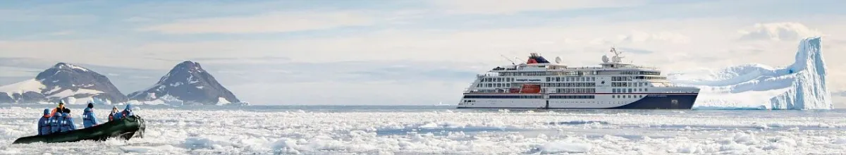 Ein Expeditionsschiff der Eisbrecherklasse von Hapag-Lloyd inmitten eines Eismeeres, vorne links ein Zodiac mit mehreren Passagieren.