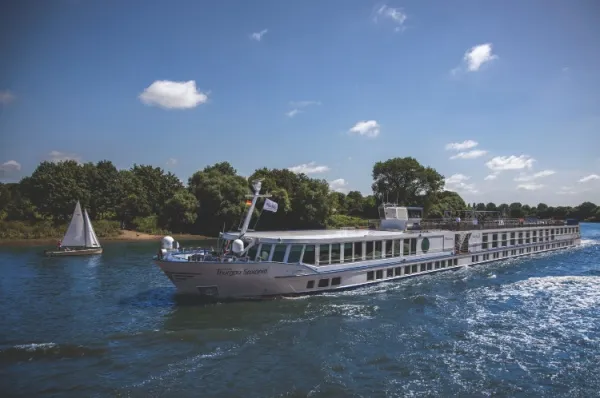 Thurgau Saxonia Die Thurgau Saxonia fährt über den Fluss, im Hintergrund ein Segelboot und grüne Waldlandschaften am Ufer.