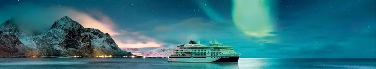 Ein beleuchtetes Hapag-Lloyd Cruises Expeditionsschiff ankert in einer Bucht vor schneebedeckten Bergen, während grüne und blaue Polarlichter den Nachthimmel erhellen –Symbolbild für unvergessliche Auszeiten, dank dem Tipp der Woche.