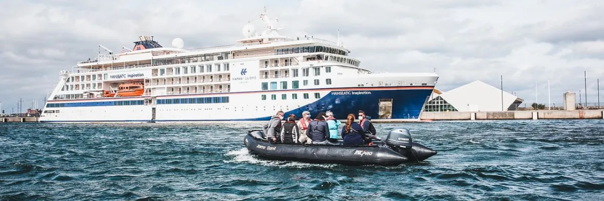 Hapag-Lloyd Cruises: HANSEATIC inspiration und Zodiac-Ausflug Das Hapag-Lloyd Expeditionskreuzfahrtschiff HANSEATIC inspiration im Hafen mit einem besetzten Zodiac-Schlauchboot im Vordergrund.