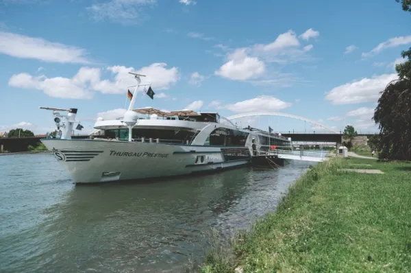 Die Thurgau Prestige angelegt am Ufer eines Flusses unter blauem Himmel. Am Ufer sieht man Wiesenlandschaft.