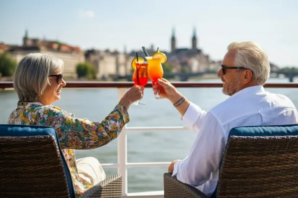Ein Mann und eine Frau sitzen auf dem Sonnendeck und stoßen mit Cocktails an während sie sich anlachen. Im Hintergrund sieht man Flusslandschaft.