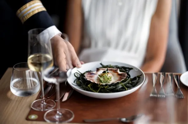 Restaurants auf der Havila Voyages Flotte Eine Frau sitzt in einem Restaurant auf der Havila Voyages Flotte und wird von einem uniformierten Kellner bedient. Sie hat ein Gericht bestellt, dass in einer Muschel serviert wird.