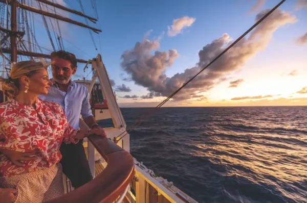 Mann und Frau stehen an Bord eines Sea Cloud Schiffes und blicken in die Ferne auf das weite Meer bei Sonnenuntergang, im Hintergrund sieht man die Segel des Schiffes.