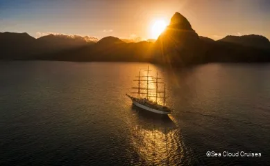 Die Sea Cloud Spirit auf dem Meer, im Hintergrund geht die Sonne hinter einer Berglandschaft unter.