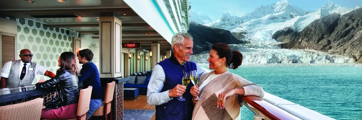 Entspannung vor den Küsten Alaskas, dank des NCL Flash Sales Ein rüstiges Pärchen an der Reling eines NCL Schiffes, links ist eine Bar mit ein paar Gästen und rechts sieht man die Küste Alaskas gesäumt von Schnee und hohen Bergen.