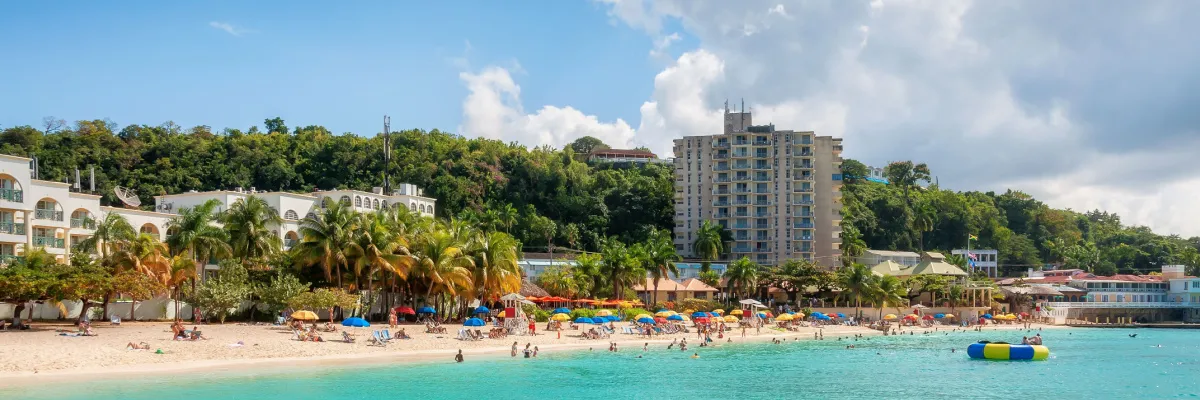 Karibik-Feeling pur mit NCL: Entspannung unter Palmen Belebter weißer Sandstrand in der Karibik mit Palmen, türkisfarbenem Wasser und Hotelanlagen im Hintergrund bei strahlendem Sonnenschein.