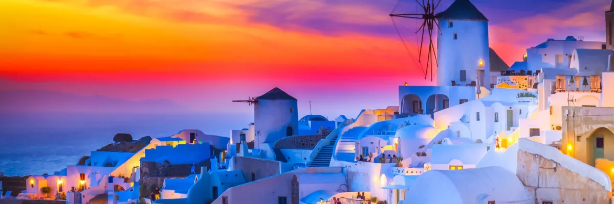 Die beleuchteten weißen Häuser und Windmühlen auf Mykonos in Griechenland vor einem farbenprächtigen Sonnenuntergang während einer NCL Kreuzfahrt.