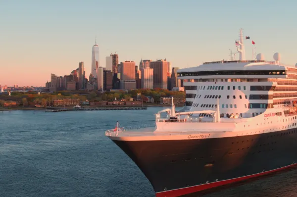 Die Queen Mary 2, wie sie New York verlässt in der Abendsonne. Im Hintergrund sind Hochhäuser.