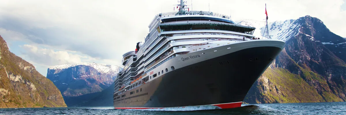 Die Queen Victoria im hohen Norden, während einer Fjordpassage. Das imposante Kreuzfahrtschiff ist von schräg vorne fotografiert und im Hintergrund sind steile Klippen und Berge zu sehen.
