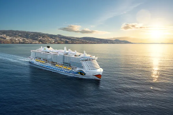 Unvergessliche Reisemomente 2026: Mit dem AIDA Sommer Bonus die schönsten Küsten der Welt entdecken Die AIDAperla fährt im goldenen Licht des Sonnenuntergangs an einer malerischen Küstenlandschaft entlang.