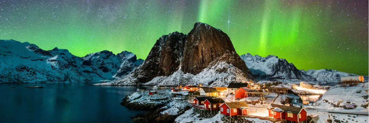 Magie des Nordens: Sichern Sie sich jetzt Ihren AIDA Sommerurlaub 2026 in Norwegen Eine malerische norwegische Fjordlandschaft bei Nacht mit traditionellen roten Hütten, schneebedeckten Bergen und leuchtend grünen Polarlichtern am Himmel – ein Symbolbild für die Faszination der AIDA Nordland-Reisen.