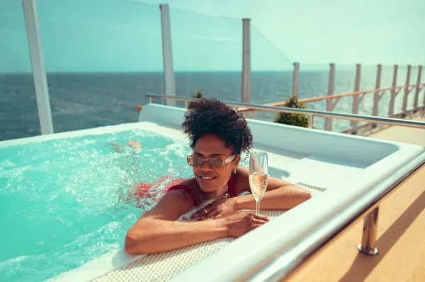 Wellness und Genuss pur: Verwenden Sie Ihr Fernweh Plus Bordguthaben für exklusive Momente im Mareel Spa Eine Frau entspannt lächelnd in einem Whirlpool auf dem Außendeck eines Cunard Schiffes und genießt ein Glas Champagner bei freier Sicht auf das offene Meer.