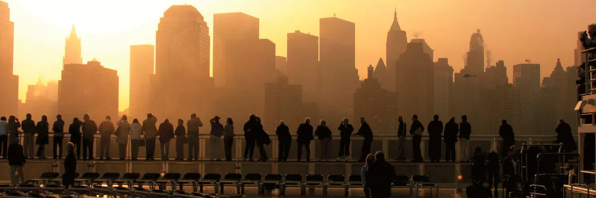 Ein magischer Moment: Die Ankunft in Manhattan bei Sonnenuntergang – ein Highlight vieler Cunard Reisen Silhouetten von Passagieren an der Reling eines Cunard Schiffes, die während der goldenen Stunde auf die dunstige Skyline von New York blicken.