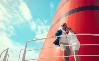 Ein glückliches älteres Paar umarmt sich lächelnd an Deck einer Cunard Queen vor dem ikonischen roten Schornstein bei sonnigem Wetter.