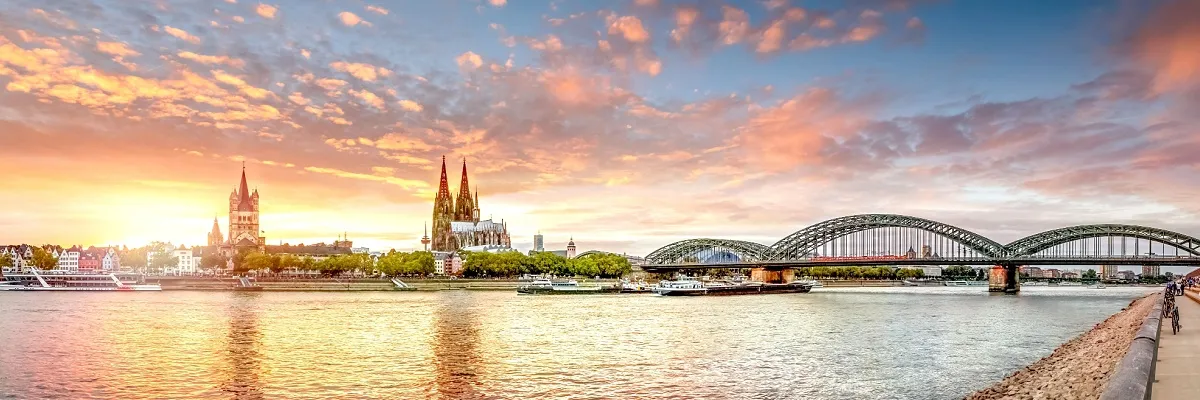 Panoramaansicht des Kölner Rheinufers im Sonnenuntergang. Zu sehen sind von links nach rechts die Kirche Groß St. Martin, der Kölner Dom und die Hohenzollernbrücke. Auf dem Rhein sind einige Schiffe zu sehen.