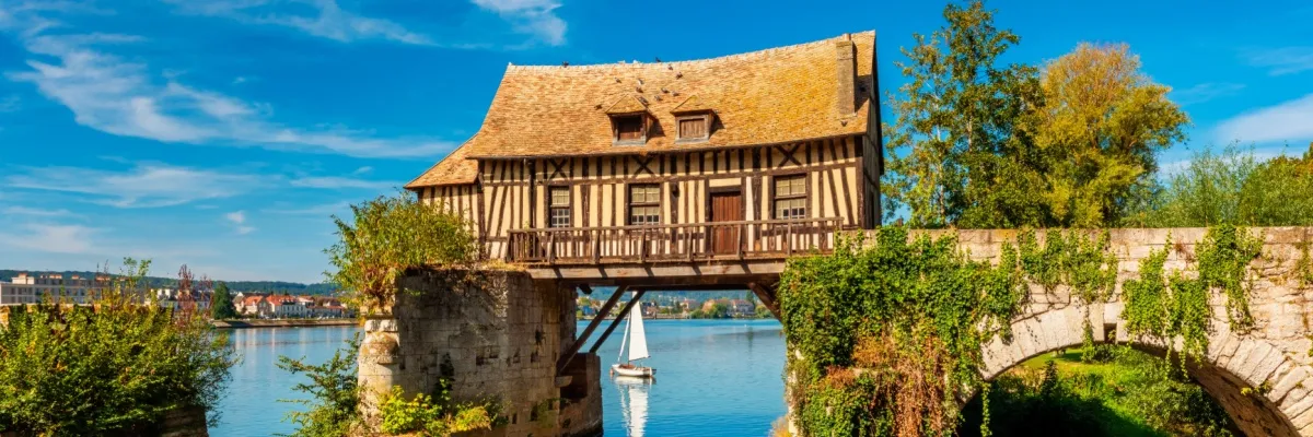 Vieux-Moulin bei Vernon. Das Fachwerkhaus der ehemaligen Wassermühle steht auf zwei Pfeilern einer mittelalterlichen Brücke über die Seine. Unterhalb des Gebäudes ist ein Segelschiff auf dem Fluss zu sehen.