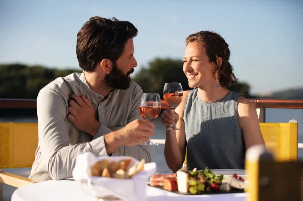Ein Paar stößt mit Gläsern Roséwein an einem elegant gedeckten Tisch auf dem Schiffdeck bei Sonnenuntergang an.