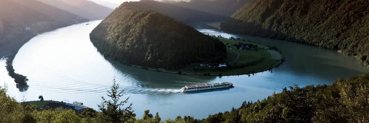 Luftaufnahme eines A-ROSA Schiffes, das durch die berühmte Schlögener Schlinge an der Donau fährt, umgeben von dicht bewaldeten Hügeln.