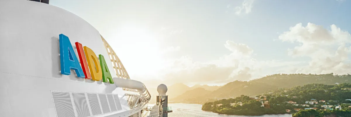 Nahaufnahme des farbenfrohen AIDA-Logos auf dem weißen Schornstein eines Kreuzfahrtschiffes vor einer sonnigen Küstenlandschaft im Hintergrund.