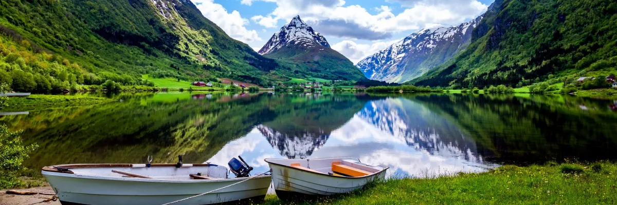 Zwei kleine weiße Ruderboote am grünen Ufer eines spiegelglatten norwegischen Fjords bei Stavanger vor schneebedeckten Berggipfeln.