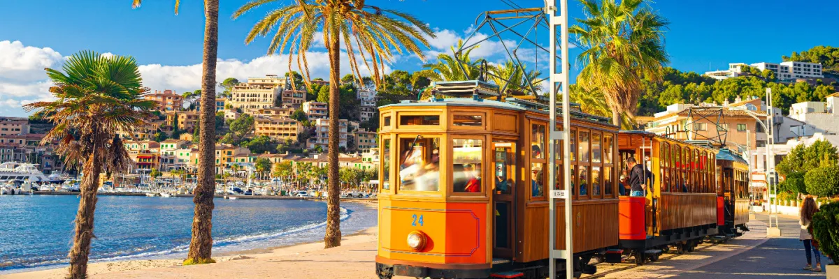 Eine historische orangefarbene Straßenbahn fährt entlang einer sonnigen Küstenpromenade mit Palmen und Blick auf eine malerische Bucht mit Booten.