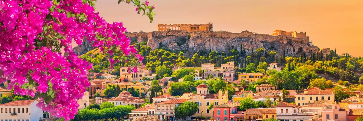 Blick auf die antike Akropolis in Athen bei Sonnenuntergang, eingerahmt von leuchtend pinkfarbenen Bougainvillea-Blüten und den Dächern der Stadt.