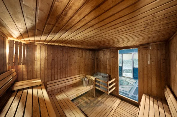 Innenansicht einer hellen Holzsauna mit mehreren Sitzreihen und einem Saunaofen in der Mitte. Im Hintergrund ist durch die offene Tür ein gefliester Raum mit Mosaikboden und einem weiteren Whirlpool oder Poolbereich zu sehen, der zum Aurea Spa gehört.