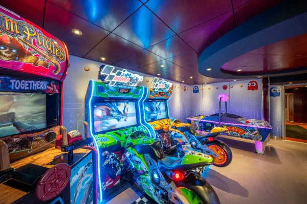 Ein bunter Spielhallenraum mit Neonlichtern an Bord der MSC Lirica. Zu sehen sind mehrere Arcade-Automaten, darunter Motorrad-Rennspiele mit physischen Motorrädern als Controller, ein Piraten-Schießspiel und ein Airhockey-Tisch im Hintergrund.