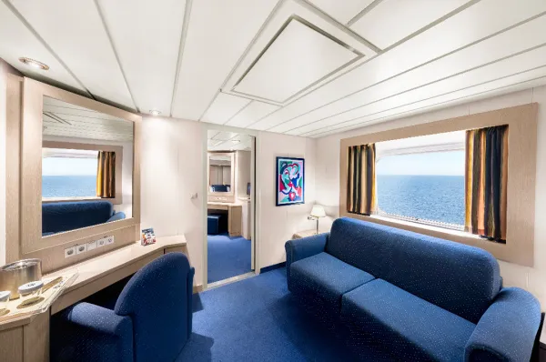 Innenansicht einer geräumigen Suite mit Meerblick auf der MSC Lirica. Die Kabine hat einen blauen Teppichboden, ein blaues Sofa unter dem großen Fenster und einen Schreibtisch mit Spiegel. Durch eine offene Tür ist ein weiterer Teil der Suite zu sehen.