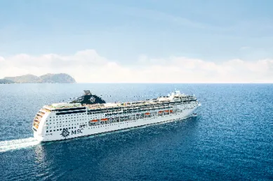 Die MSC Lirica, ein weißes Kreuzfahrtschiff mit dem MSC-Logo am Schornstein, fährt auf blauem Meer. Am Horizont sind grüne Inseln und ein klarer Himmel zu sehen, während das Schiff eine leichte Gischt hinter sich herzieht.