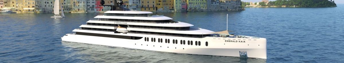 Die Emerald Kaia in Seitenansicht auf See vor einer kleinen Küstenstadt mit bunten Häusern.