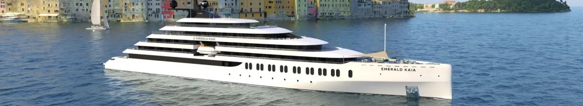 Die Emerald Kaia in Seitenansicht auf See vor einer kleinen Küstenstadt mit bunten Häusern.