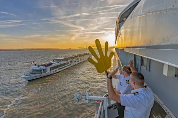 Zwei Crew-Mitglieder winken von der Scenic Eclipse zur Scenic Gem, die an ihnen vorbeischippert, im Hintergrund geht die Sonne unter.