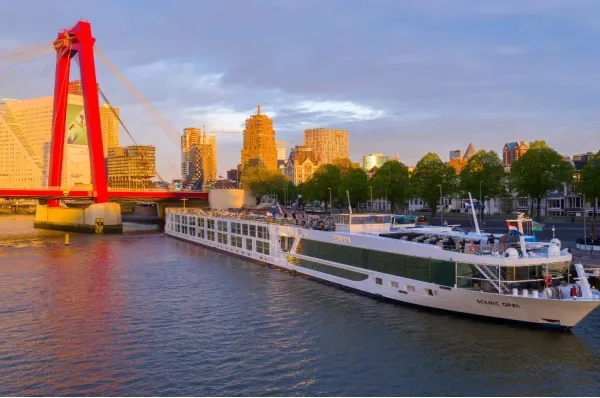 Das Flussschiff Scenic Opal vor Rotterdam mit roter Brücke und Hochhäusern im Hintergrund.
