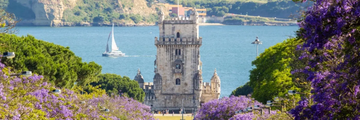 Mit Swan Hellenic nach Europa Der Tturm von Belém in Lissabon an einem sommerlichen Tag. schiffe segeln im Kanal und die Umgebung ist von lila Blüten eingerahmt.